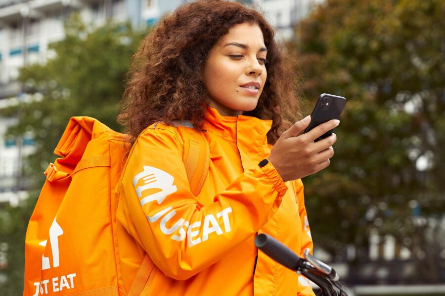 En kvinna i jacka och ryggsäck med texten Just Eat, kollar sin mobil